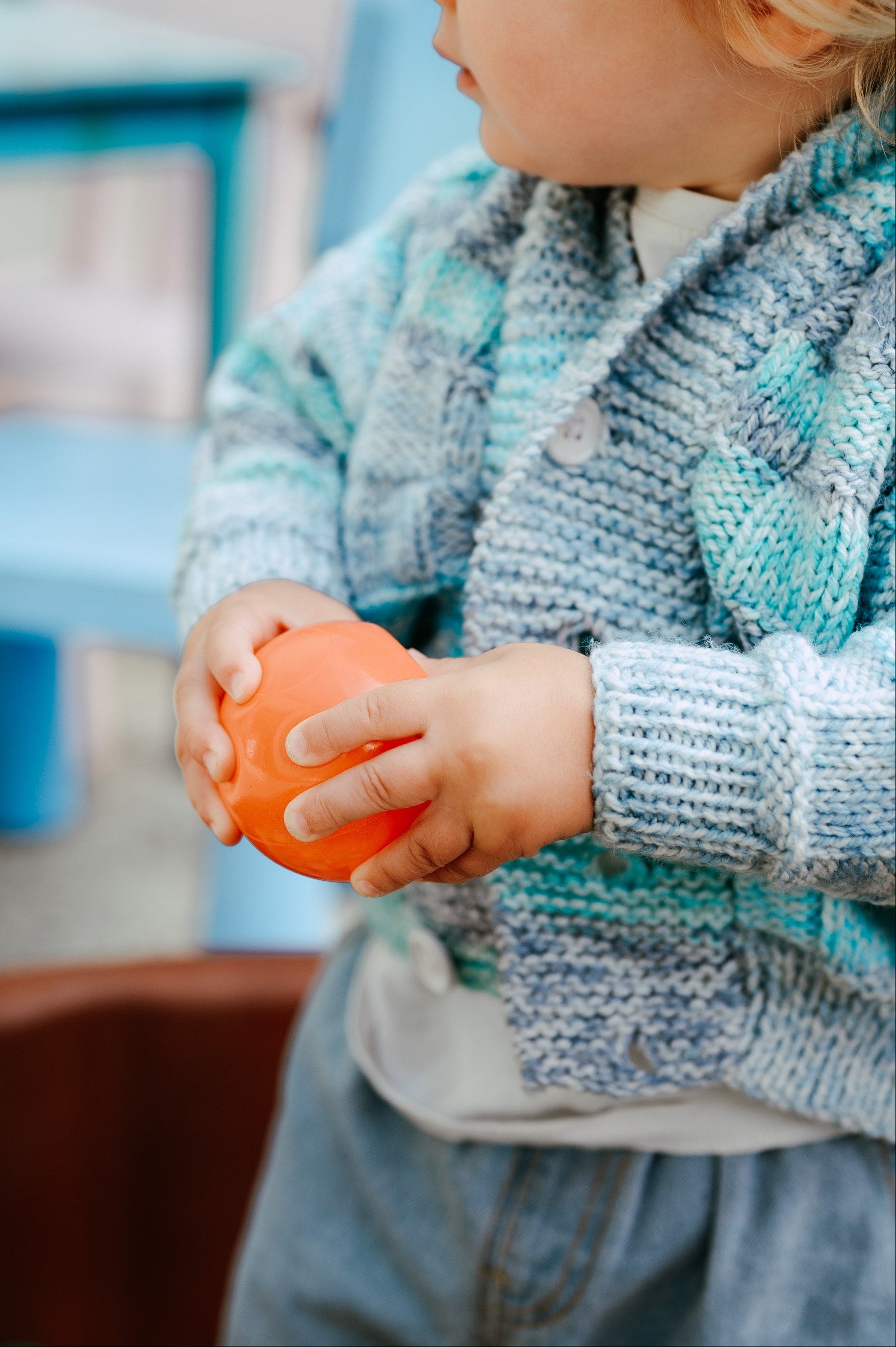 Child holding an orange ball outdoors with blurred furniture in the background