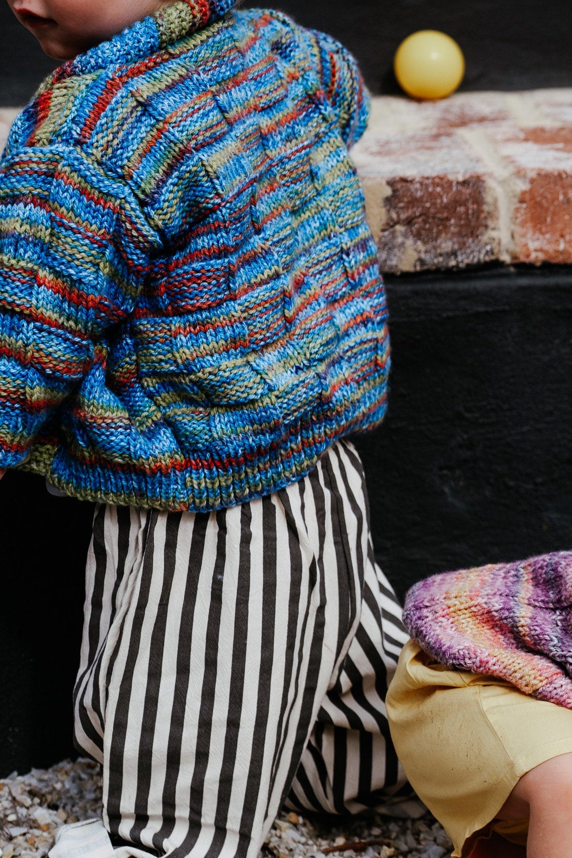 Children playing outdoors in striped knitted sweaters with balls on a stone ledge.