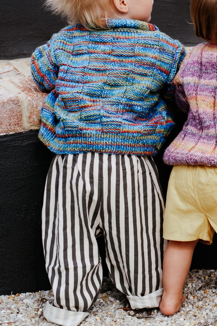 Two children wearing colorful knitted sweaters, one in striped pants and one in yellow shorts, standing on a gravel surface.