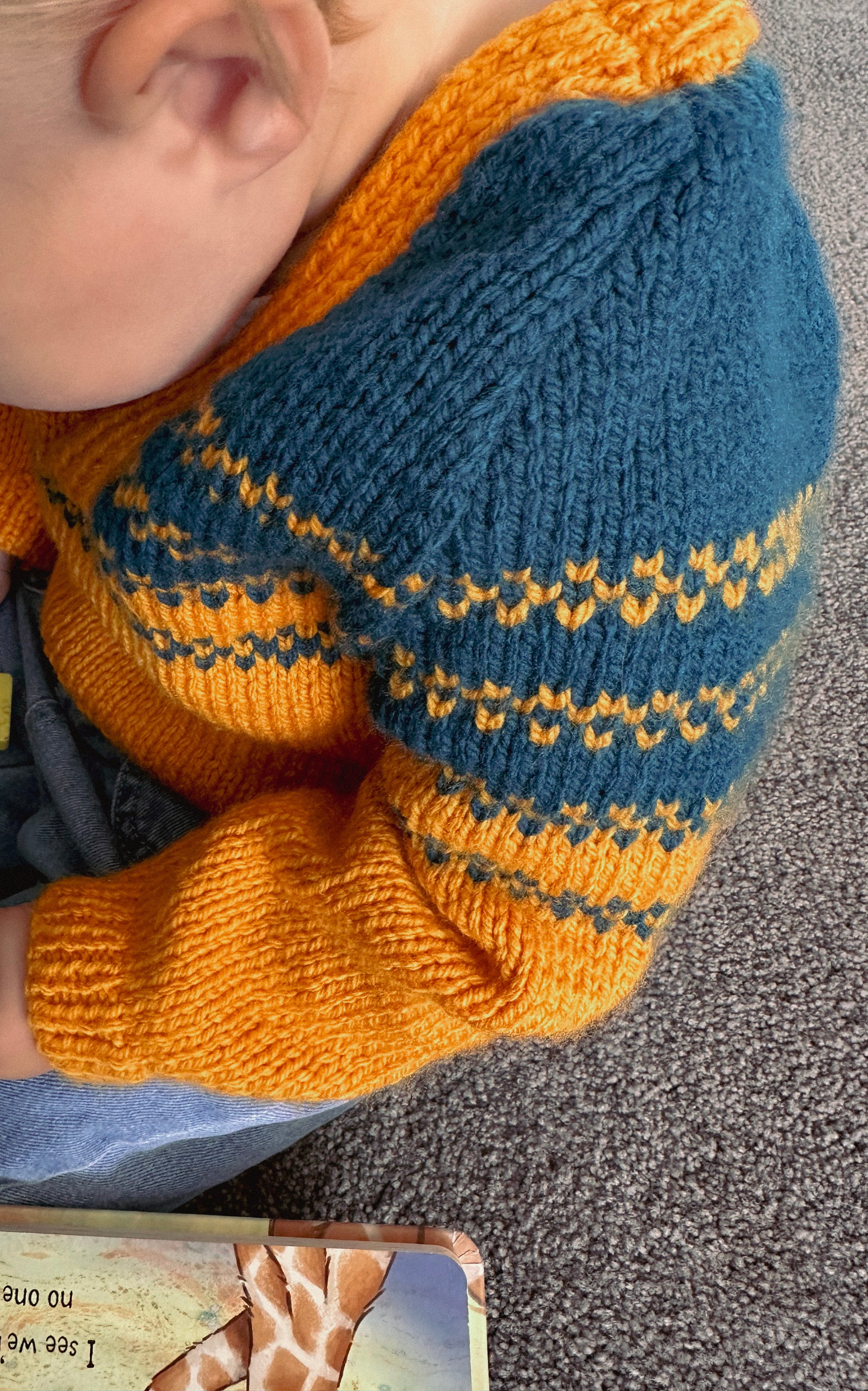 A close-up of a child wearing a hand-knit sweater with a blue and mustard yellow pattern, sitting on a gray carpet.