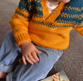 A close-up of a child wearing a hand-knit V-neck sweater with a mustard yellow base and dark teal geometric patterns, paired with light blue jeans.