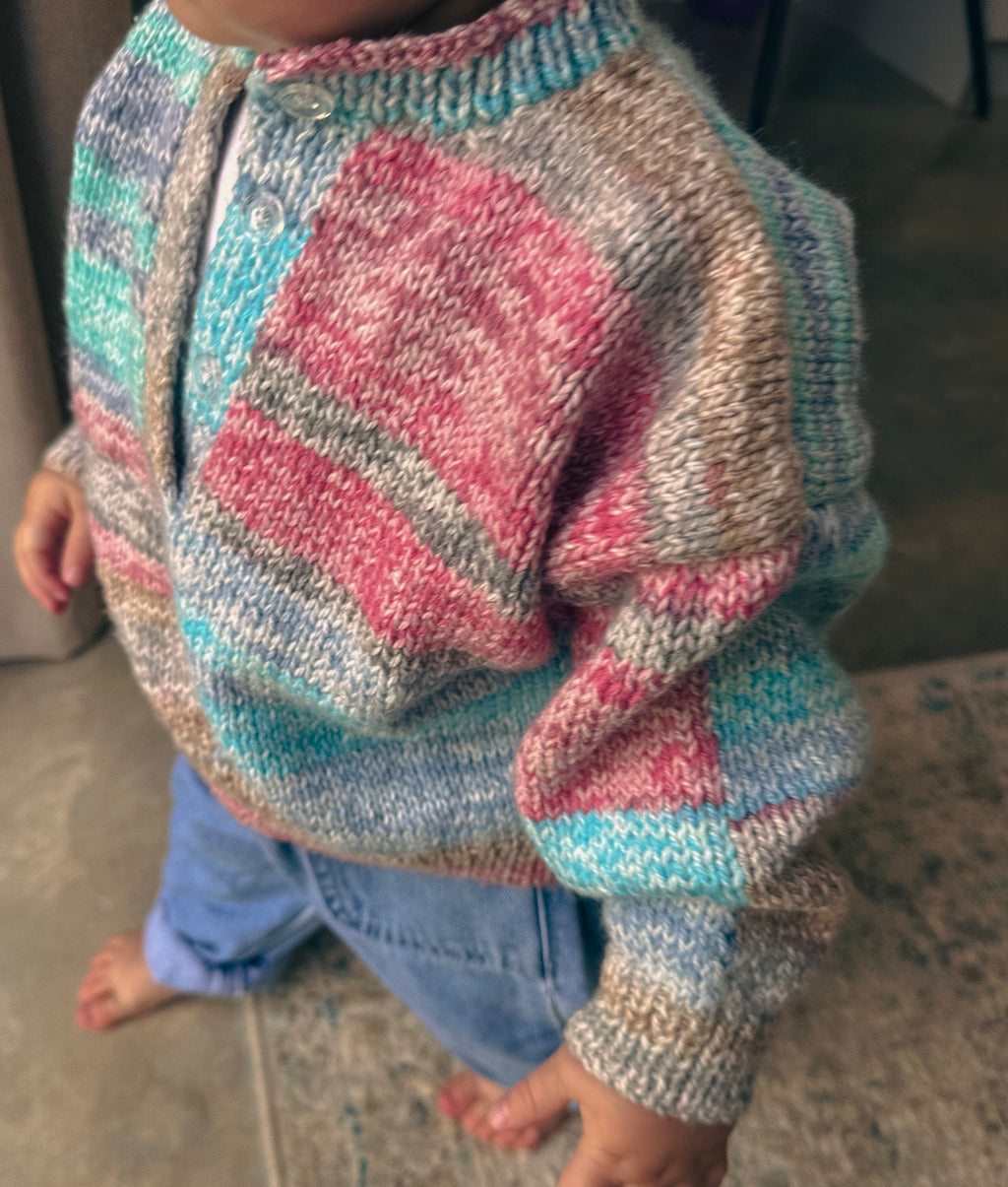 A close-up of a child wearing a hand-knit cardigan sweater with a multicolor striped pattern of pink, blue, gray, and beige, paired with light blue jeans.