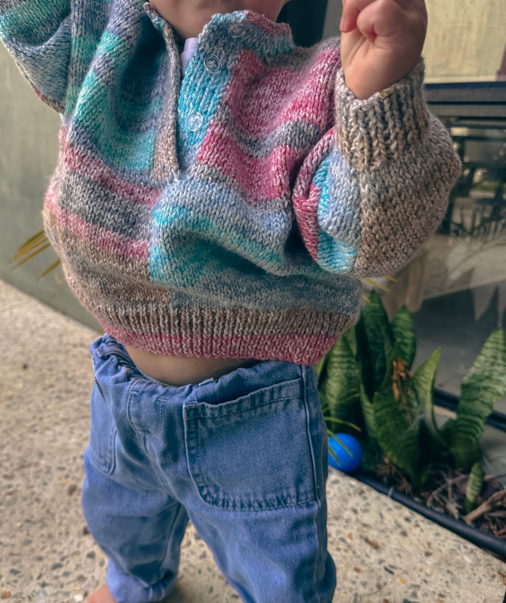 A close-up of a child wearing a hand-knit half-button sweater with wavy stripes of pink, teal, blue, and gray, paired with blue jeans.