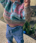 A close-up of a child wearing a hand-knit half-button sweater with wavy stripes of pink, teal, blue, and gray, paired with blue jeans.