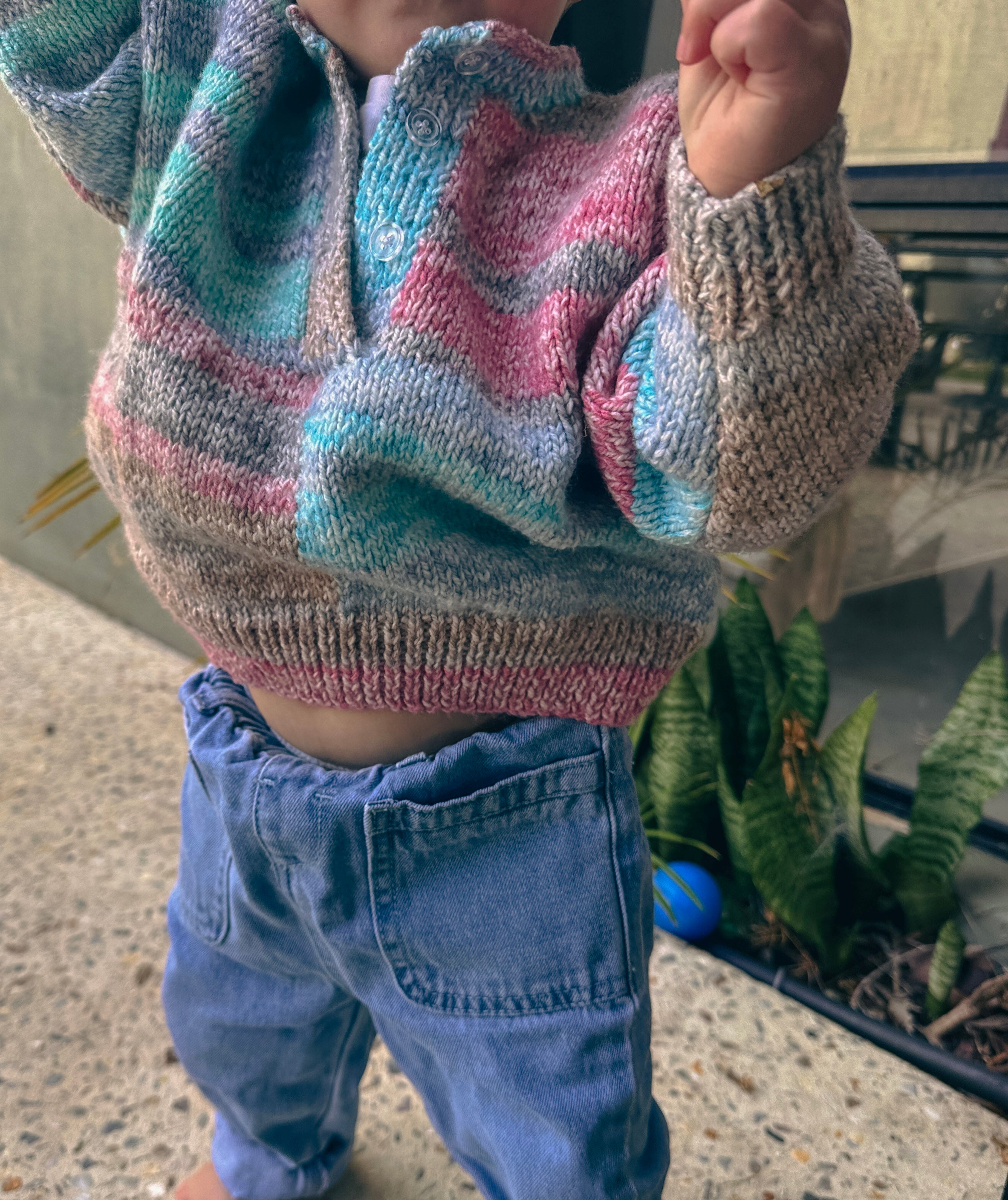 A close-up of a child wearing a hand-knit half-button sweater with wavy stripes of pink, teal, blue, and gray, paired with blue jeans.
