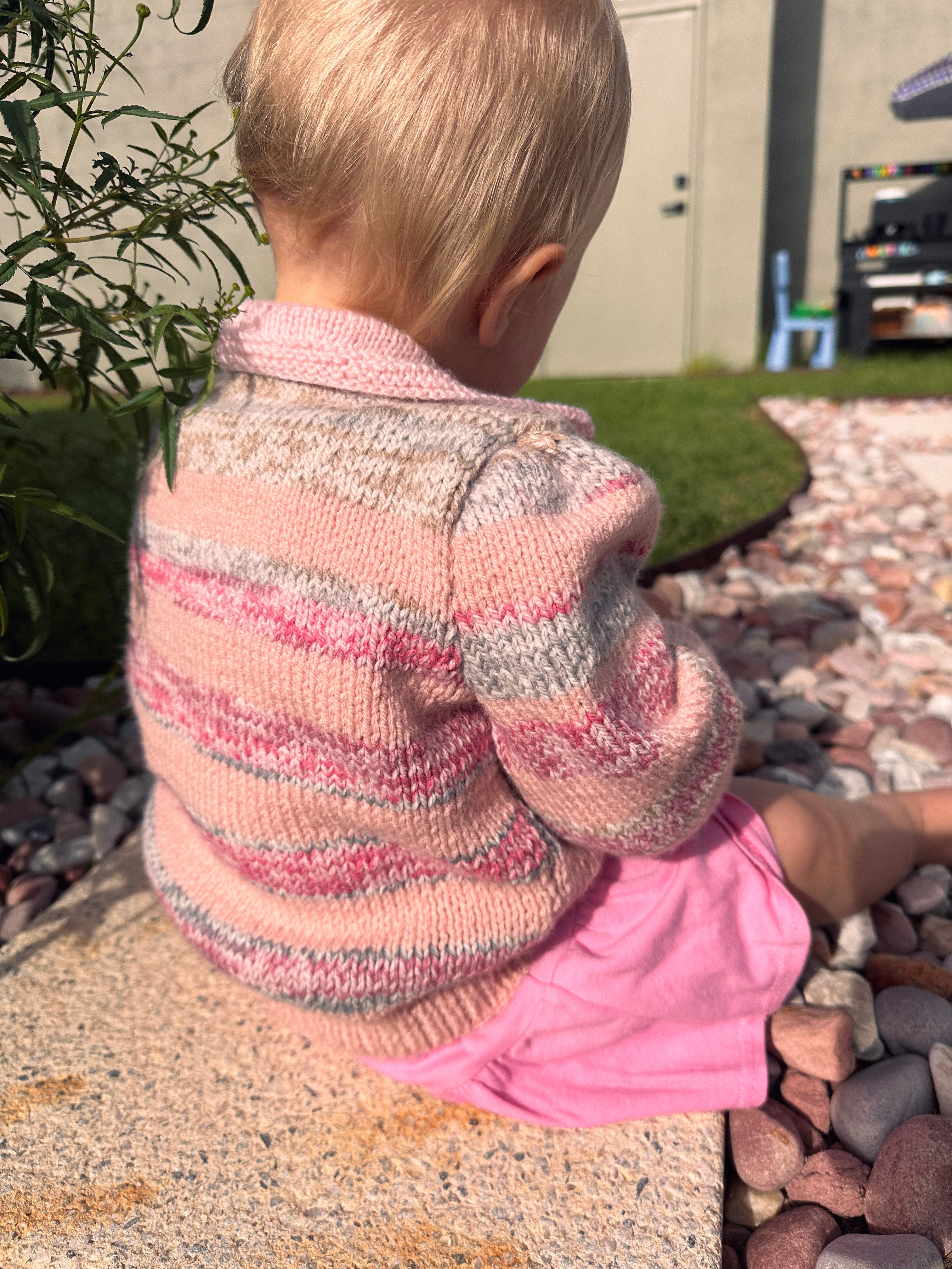 A toddler sitting outdoors on a stone ledge, viewed from behind, wearing a pink and beige striped knit cardigan and a pink skirt.