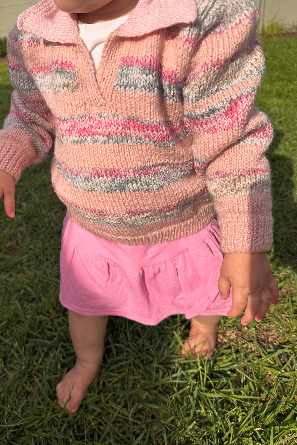 A close-up of a child outdoors on grass, wearing a pink knit long-sleeve sweater with horizontal multicolor stripes and a pink skirt.