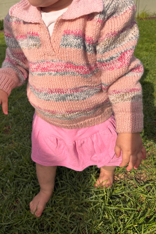 A close-up of a child outdoors on grass, wearing a pink knit long-sleeve sweater with horizontal multicolor stripes and a pink skirt.