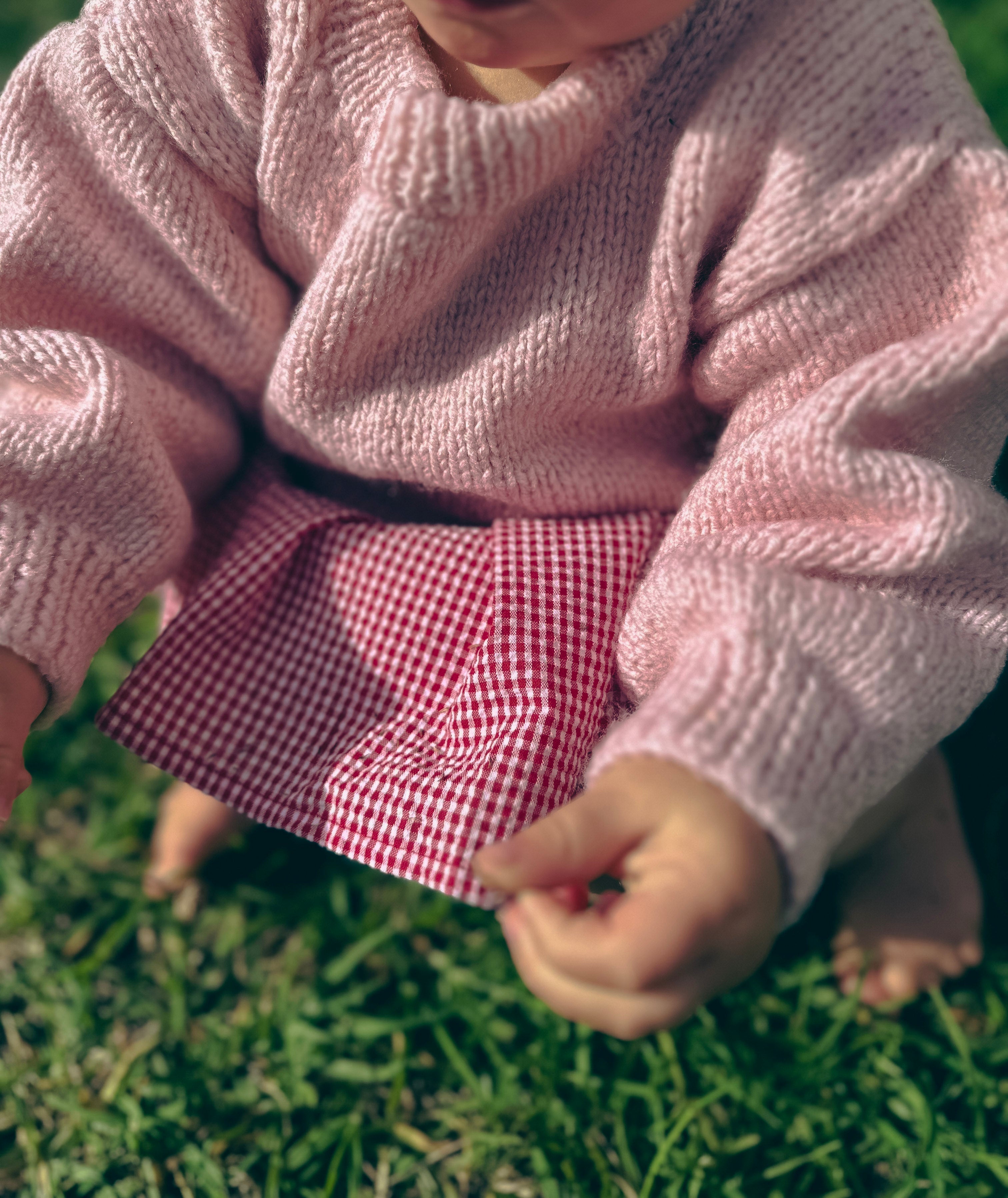 A close-up lifestyle shot of a baby wearing a pink knit sweater and a red-and-white gingham skirt, sitting on green grass outdoors.
