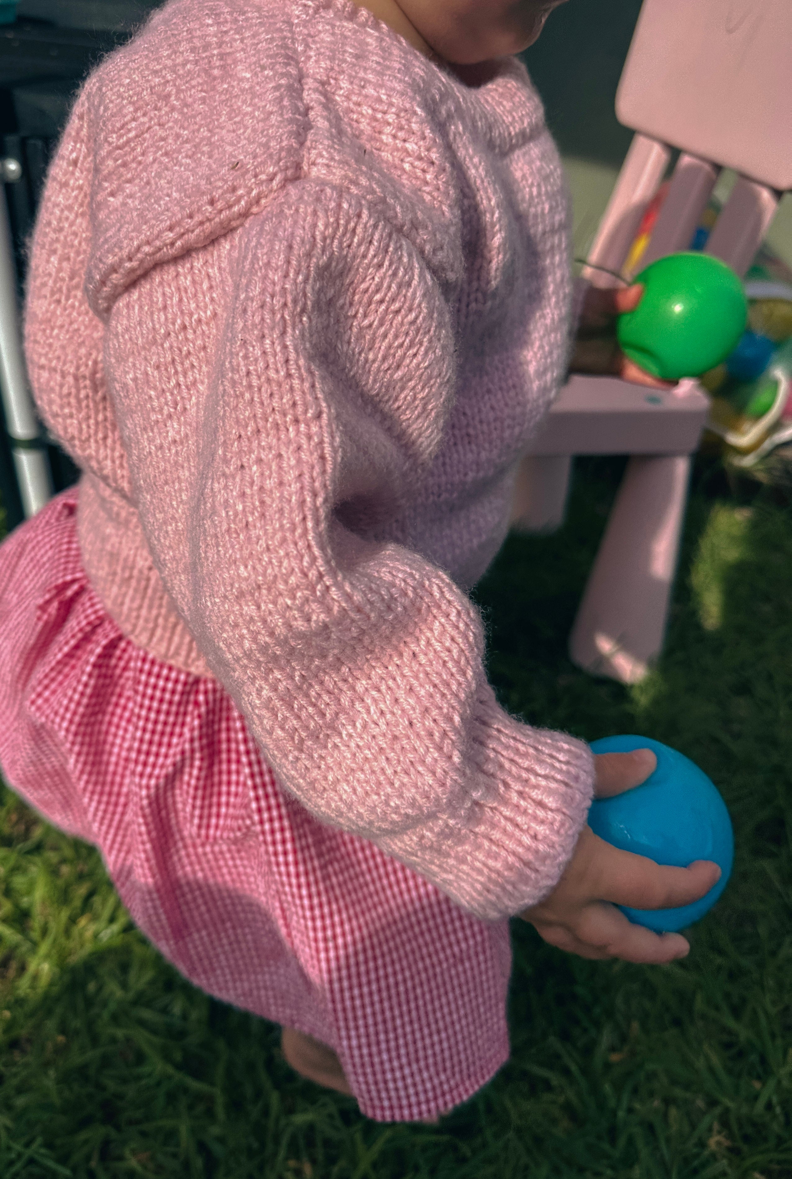 A child wearing a chunky knit light pink sweater and a pink and white gingham skirt, holding a blue ball on a grassy outdoor surface.