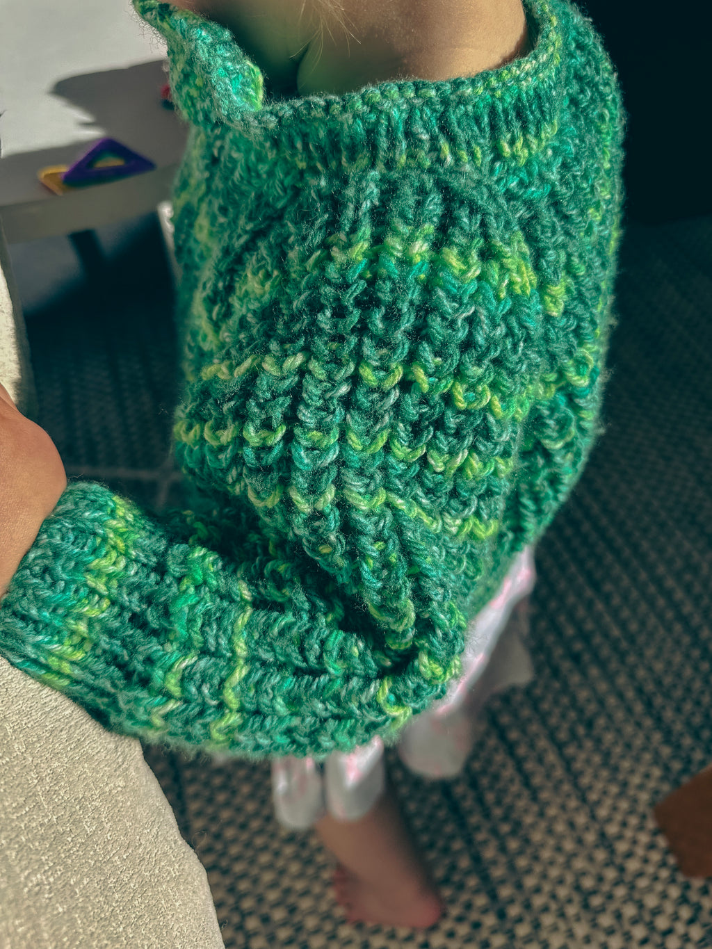 A close-up of a child wearing a chunky knit sweater in variegated green and yellow yarn, with a textured cable-knit pattern and ribbed cuffs.