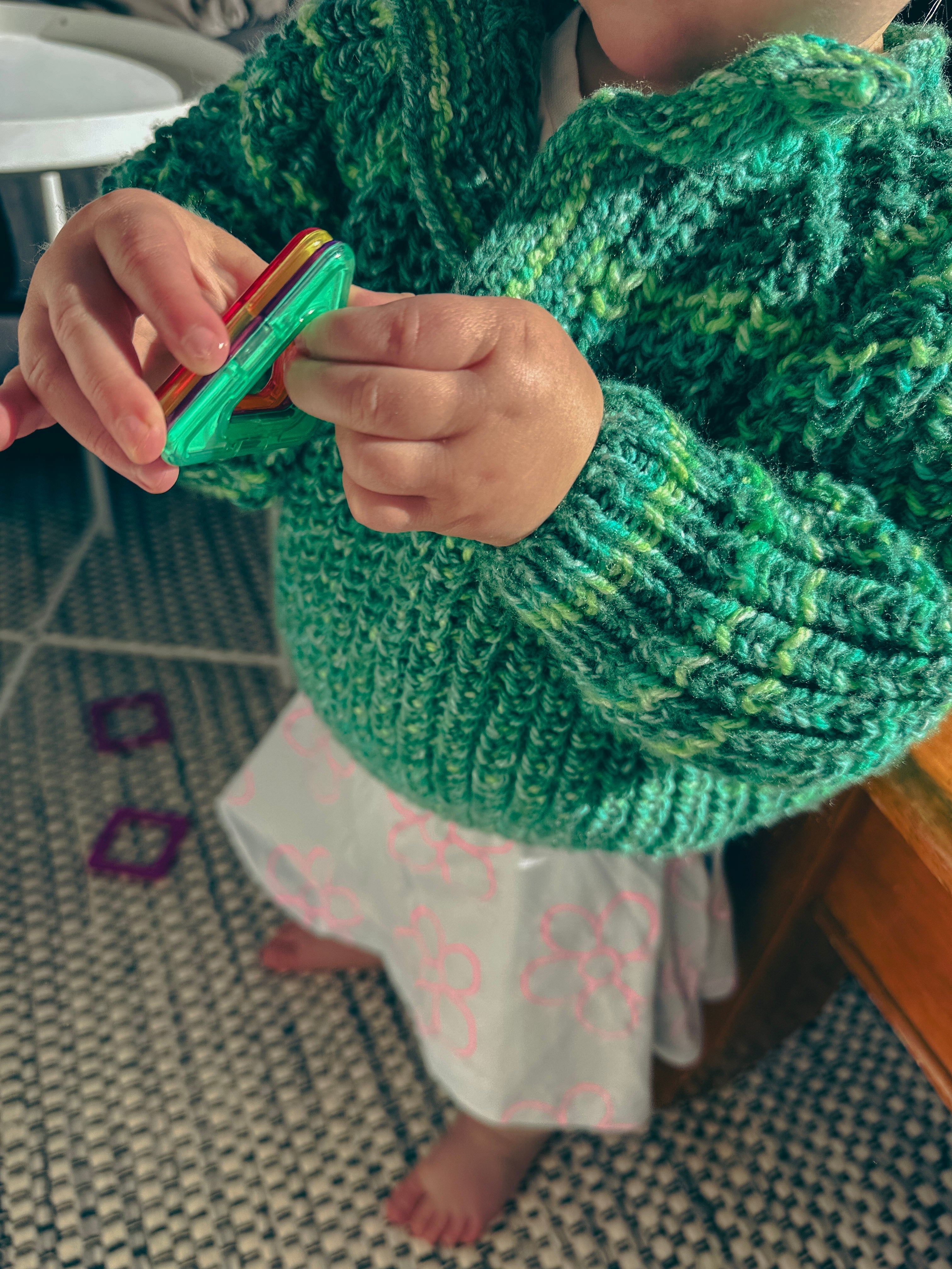 A close-up of a child wearing a chunky green and yellow variegated knit sweater and a white skirt with pink patterns, holding a small colorful toy in a home setting.