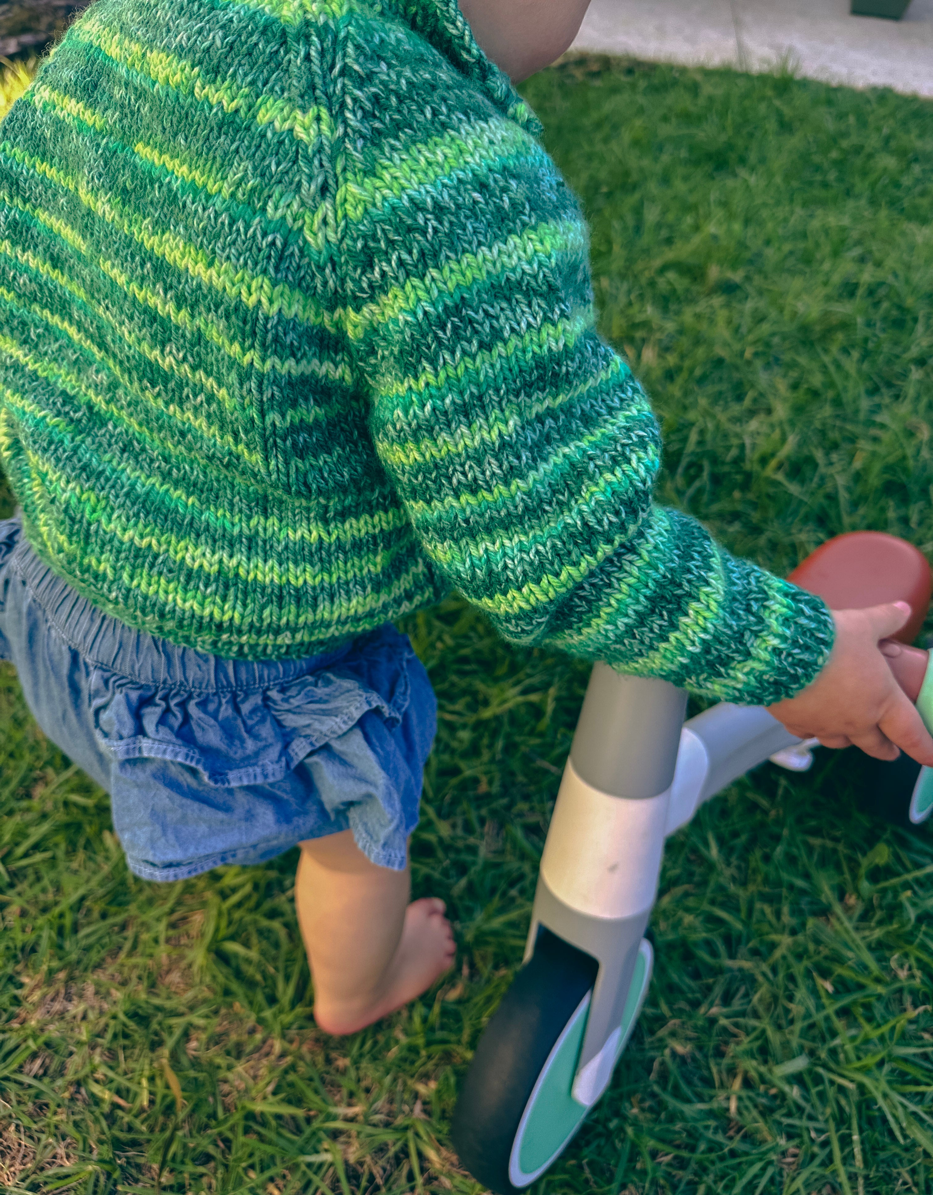 A child outdoors on grass wearing a green and yellow striped knit sweater and denim shorts, standing beside a small balance toy.