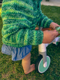A close-up outdoor shot of a child wearing a green and yellow striped chunky knit sweater and denim shorts, standing on grass next to a tricycle.