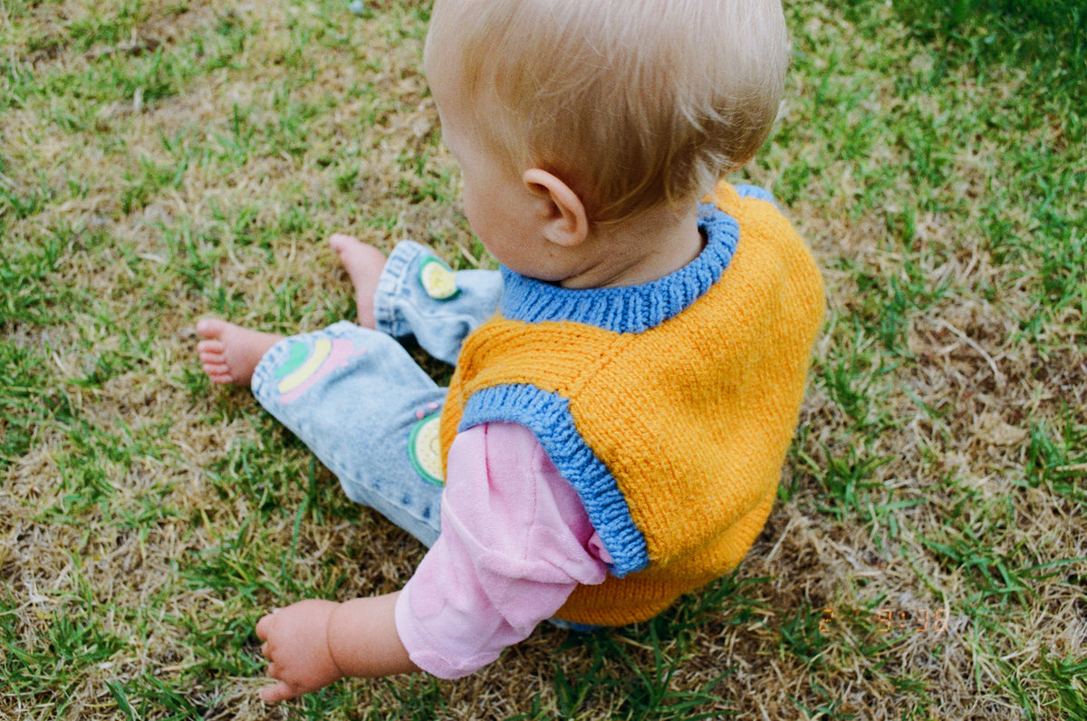 Baby sitting on grass wearing a colorful sweater and jeans.