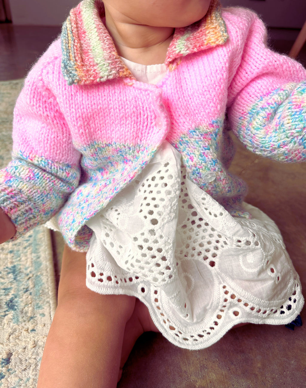 Child wearing a pink knitted sweater with colorful trim sitting on a carpeted floor.