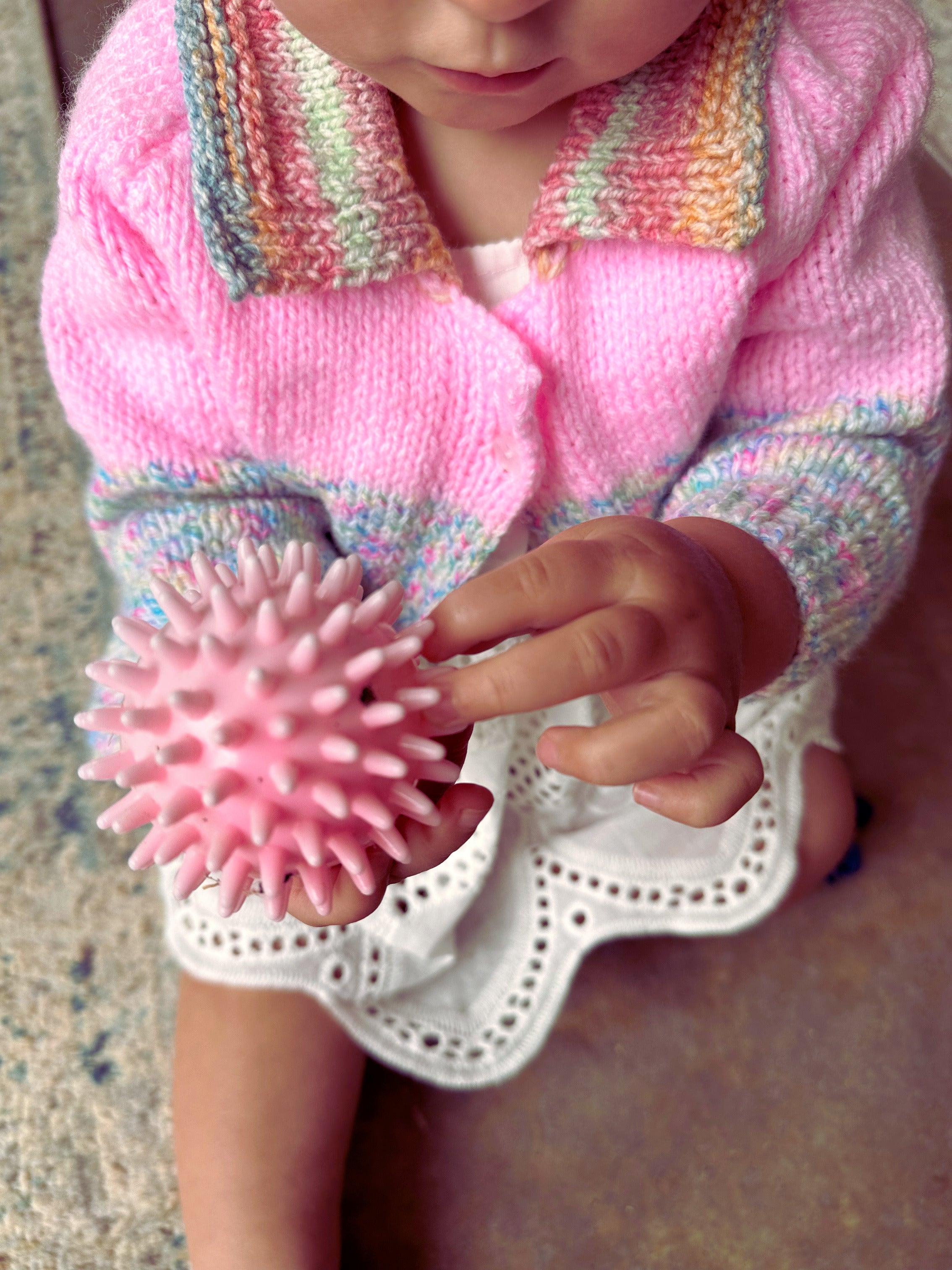 Child holding a pink textured ball wearing a colorful knitted sweater.
