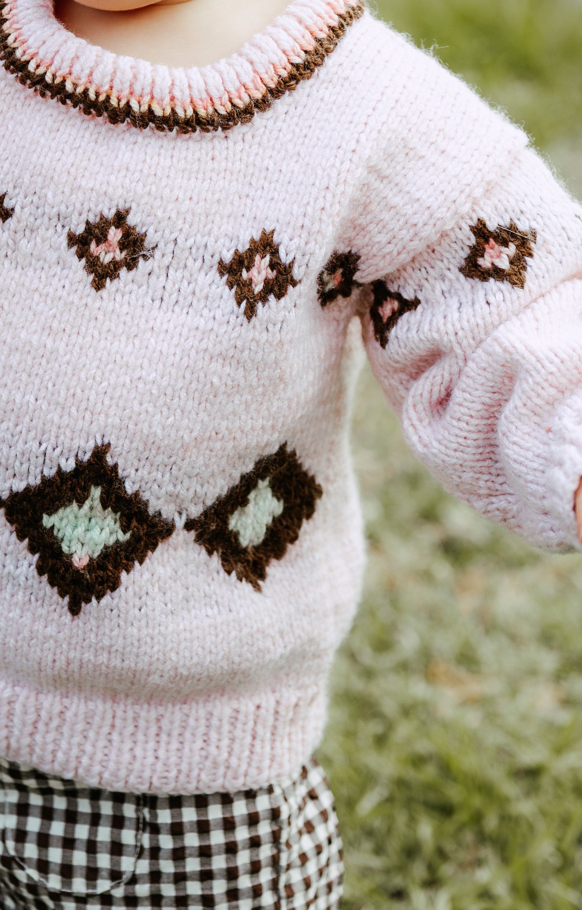 A close-up of a baby wearing a light pink hand-knit sweater with brown and white geometric patterns, paired with brown and white gingham shorts, standing outdoors on grass.
