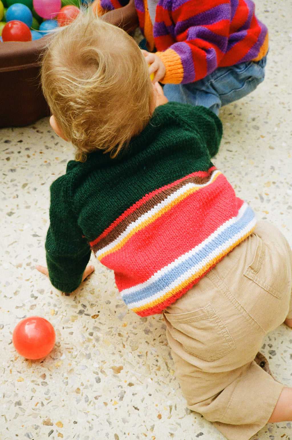 A baby crawling on a speckled floor wearing a green knit sweater with red, orange, yellow, blue, and white stripes around the hem, paired with tan pants. Another child in a colorful striped sweater is partially visible in the background near a ball pit.