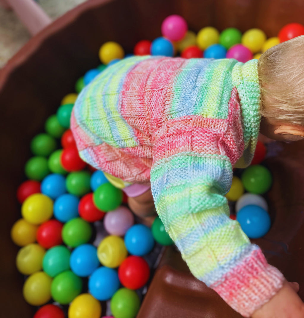 Child's hand wearing a colorful striped glove reaching into a bowl filled with colorful balls.
