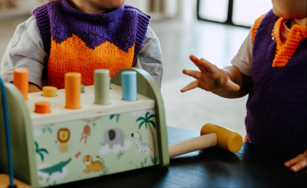 two toddlers wearing purple and orange knit vest an checked pants playing with toys