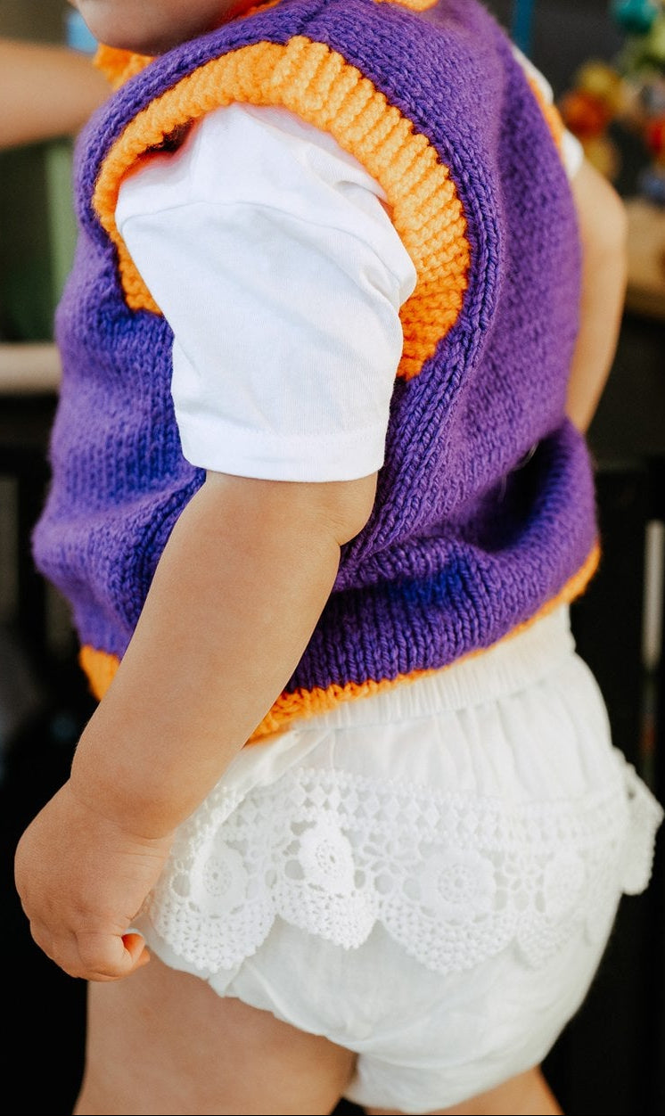 Child wearing a purple and orange vest with white romper in a casual setting
