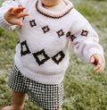 A baby wearing a light pink hand-knit sweater with brown and white diamond patterns, paired with brown and white gingham shorts, standing outdoors on grass.