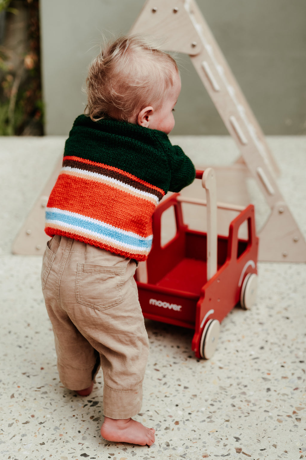 A toddler standing barefoot outdoors next to a red wooden toy truck, wearing a colorful striped knit sweater and beige pants.