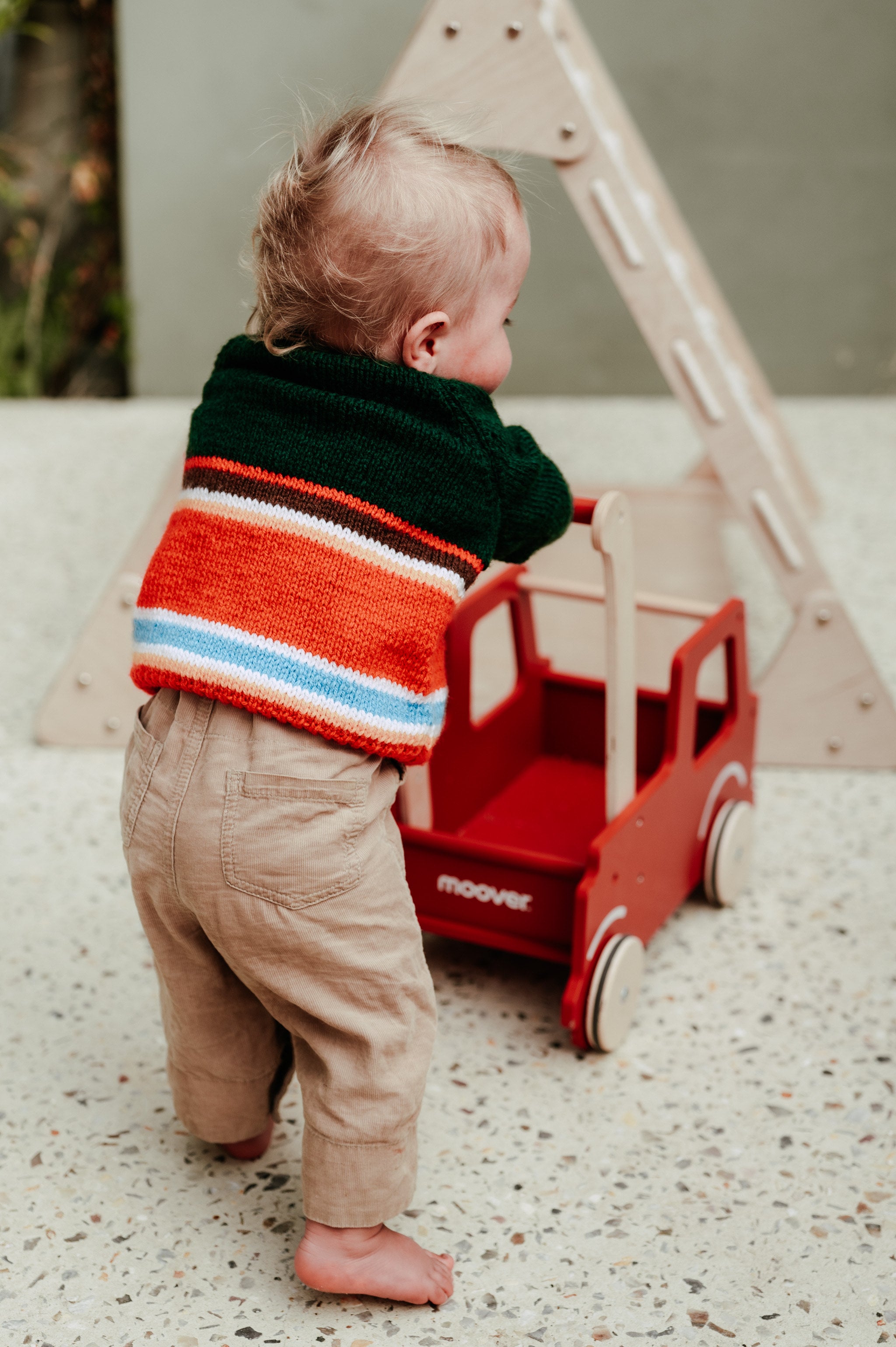 A toddler standing barefoot outdoors next to a red wooden toy truck, wearing a colorful striped knit sweater and beige pants.