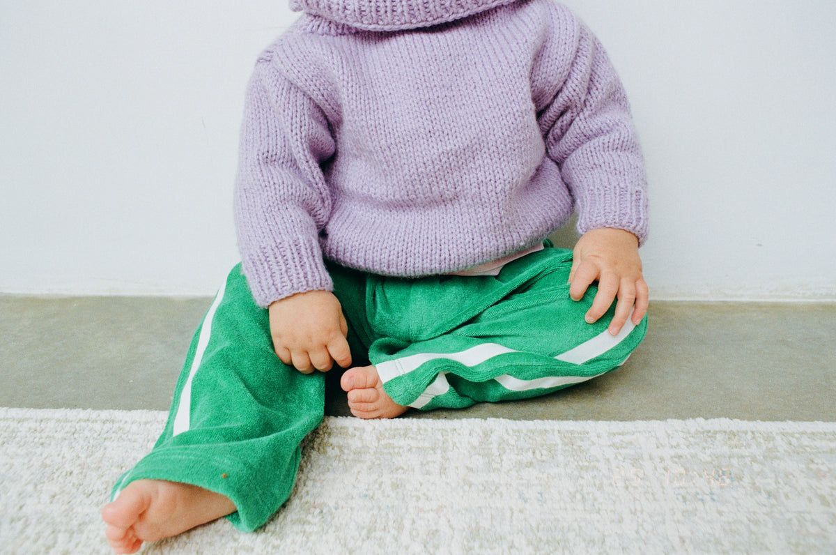 Child wearing a purple sweater and green pants sitting on a white surface.