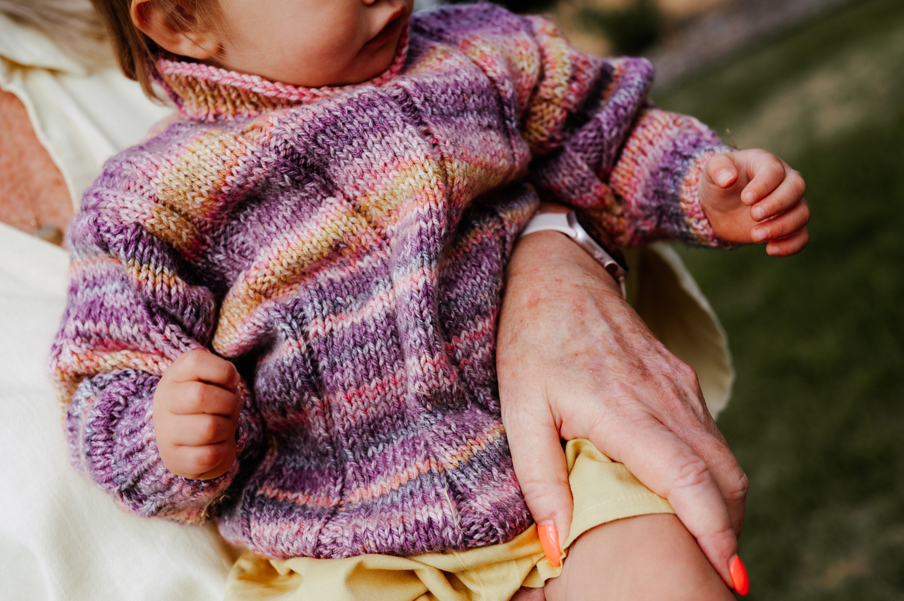 Baby wearing a colorful striped sweater held by an adult outdoors.