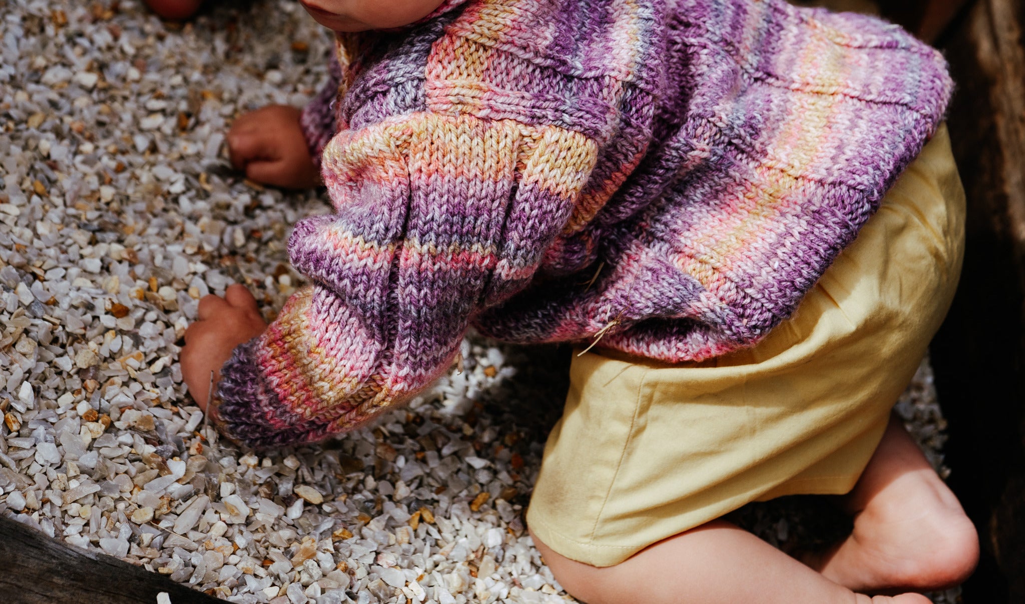 Close-up of a child's legs wearing handmade knitted purple sweater colorful striped socks and yellow pants on a gravel surface.